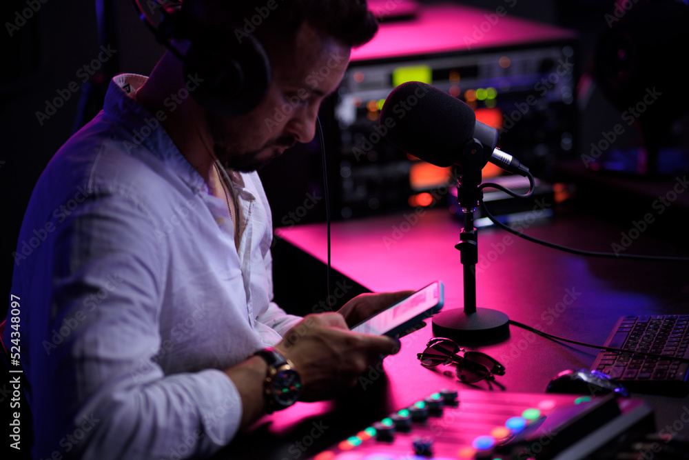 Young man host enjoying podcasting in studio, speaking into a microphone, holding a phone, using laptop. Handsome podcaster streaming live audio podcast Young man host enjoying podcasting in studio, speaking into a microphone, holding a phone, using laptop. Handsome podcaster streaming live audio podcast
