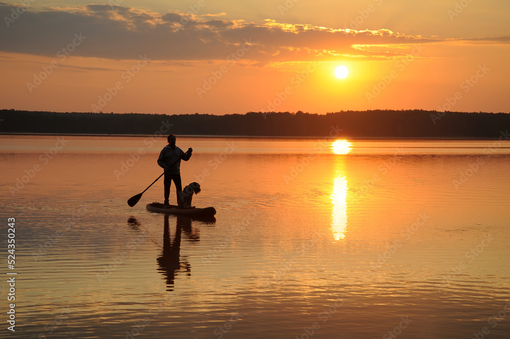 Silhouettes of a man with a dog paddling in lake water on a SUP board at sunset. Vacation, tourism, active lifestyle, hobbies.