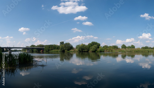 Rzeka Odra, w tle most lekko pochmurna pogoda, błękit nieba zieleń przy brzegu,  pora letnia, Odra w odcinku województwa Opolskiego