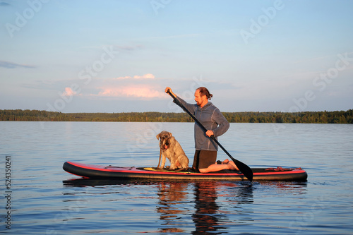 A mature man with a dog is paddling on the lake water on a SUP board. Vacation, tourism, active lifestyle, hobbies.