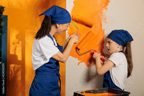Small family painting walls with orange paint to redecorate apartment space with tools and paintbrush. Woman with little girl renovating house room with roller brush and color, teamwork improvement.