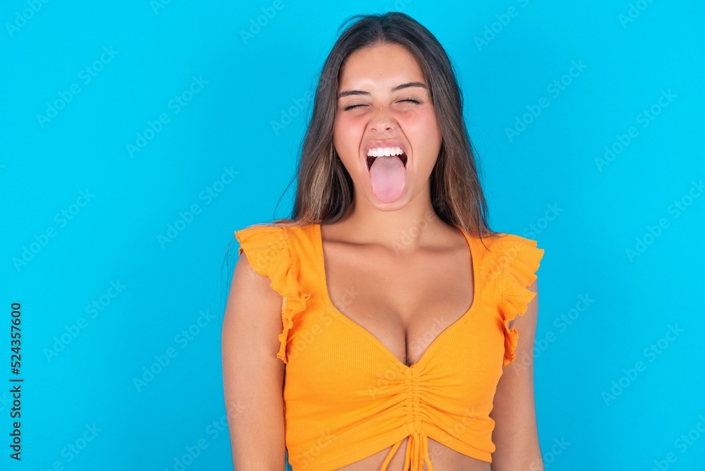 beautiful brunette woman wearing orange tank top over blue background with happy and funny face smiling and showing tongue. beautiful brunette woman wearing orange tank top over blue background with happy and funny face smiling and showing tongue.