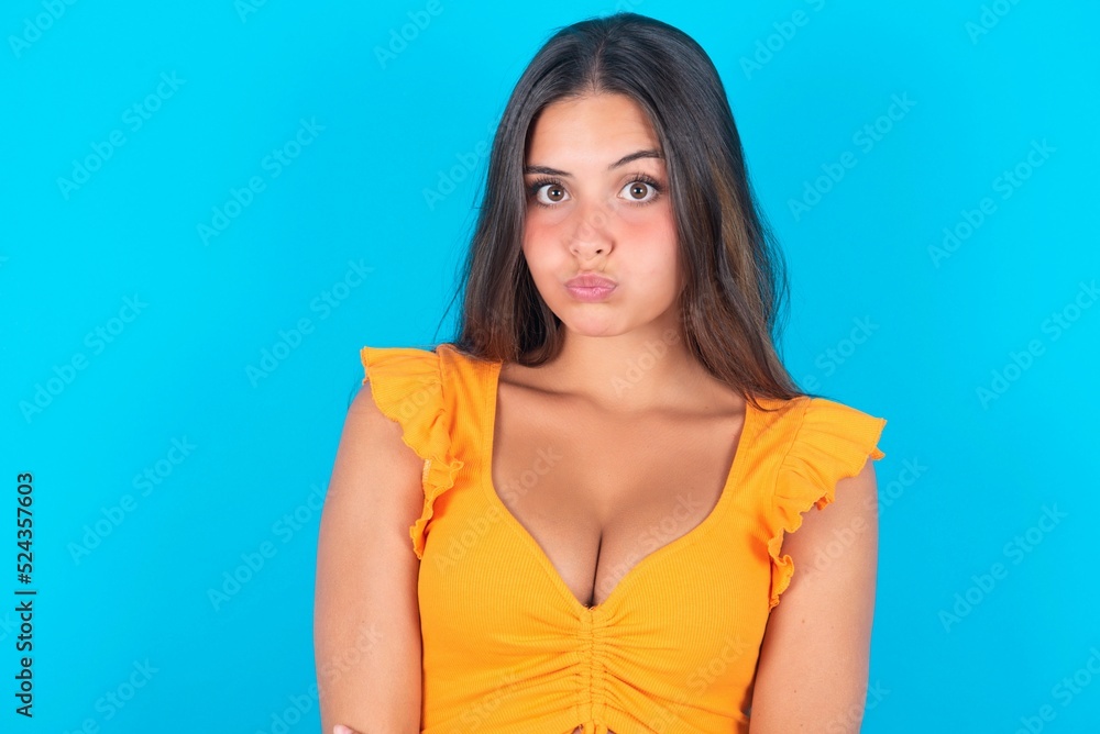 beautiful brunette woman wearing orange tank top over blue background puffing cheeks with funny face. Mouth inflated with air, crazy expression. beautiful brunette woman wearing orange tank top over blue background puffing cheeks with funny face. Mouth inflated with air, crazy expression.