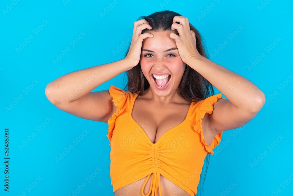 Shocked panic brunette woman wearing orange tank top over blue background  holding hands on head and screaming in despair and frustration.