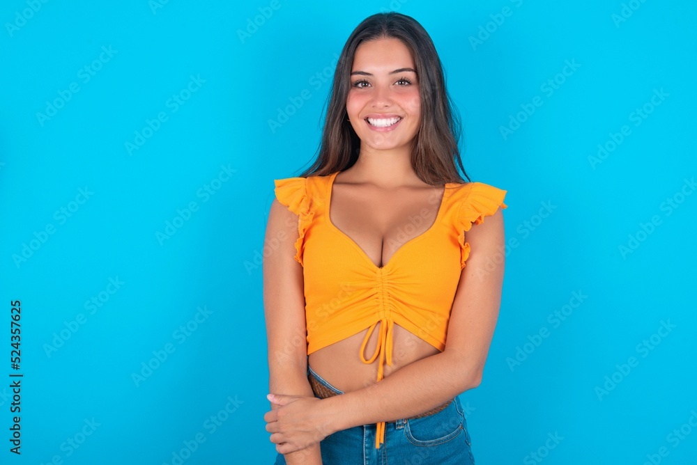 brunette woman wearing orange tank top over blue background  with broad smile, shows white teeth, feeling confident rejoices having day off.