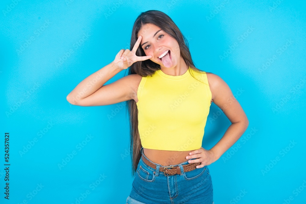 brunette woman wearing yellow tank top over blue background making v-sign near eyes. Leisure, coquettish, celebration, and flirt concept. brunette woman wearing yellow tank top over blue background making v-sign near eyes. Leisure, coquettish, celebration, and flirt concept.