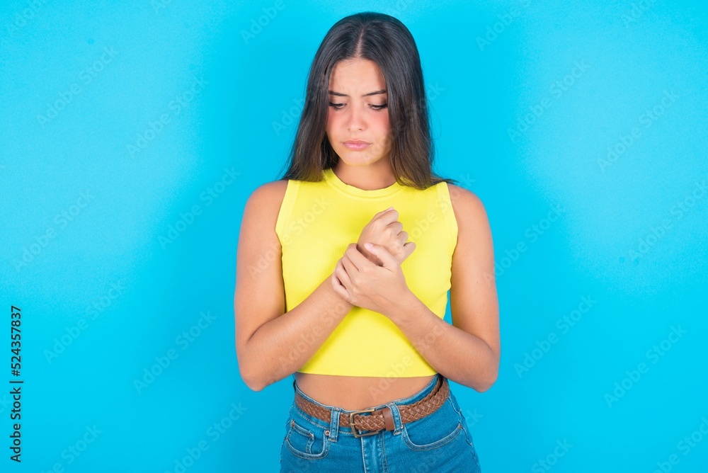 Sad brunette woman wearing yellow tank top over blue background feeling upset while spending time at home alone staring at camera with unhappy or regretful look. Sad brunette woman wearing yellow tank top over blue background feeling upset while spending time at home alone staring at camera with unhappy or regretful look.