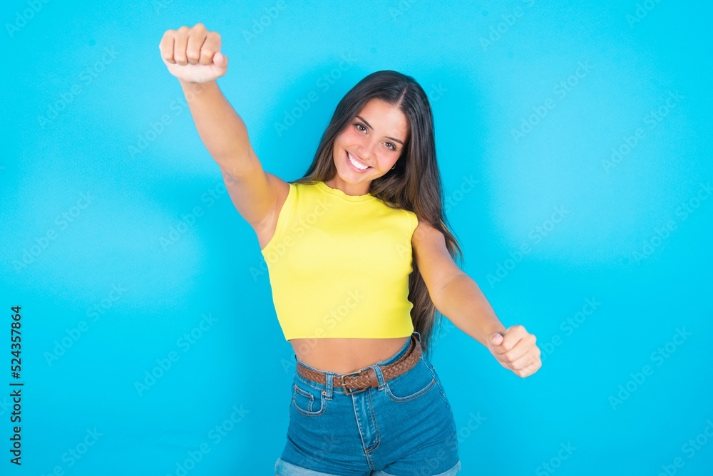 Portrait of charming brunette woman wearing yellow tank top over blue background, smiling broadly while holding hands over her head. Confidence and relax concept. Portrait of charming brunette woman wearing yellow tank top over blue background, smiling broadly while holding hands over her head. Confidence and relax concept.