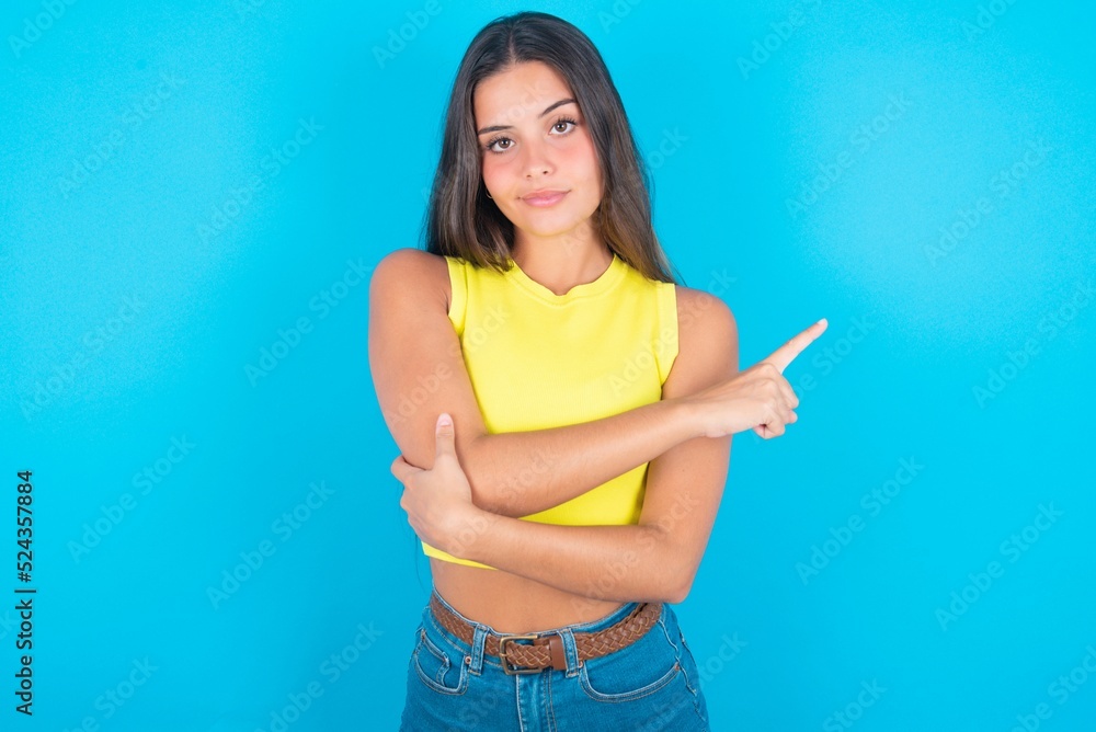 brunette woman wearing yellow tank top over blue background smiling broadly at camera, pointing fingers away, showing something interesting and exciting. brunette woman wearing yellow tank top over blue background smiling broadly at camera, pointing fingers away, showing something interesting and exciting.