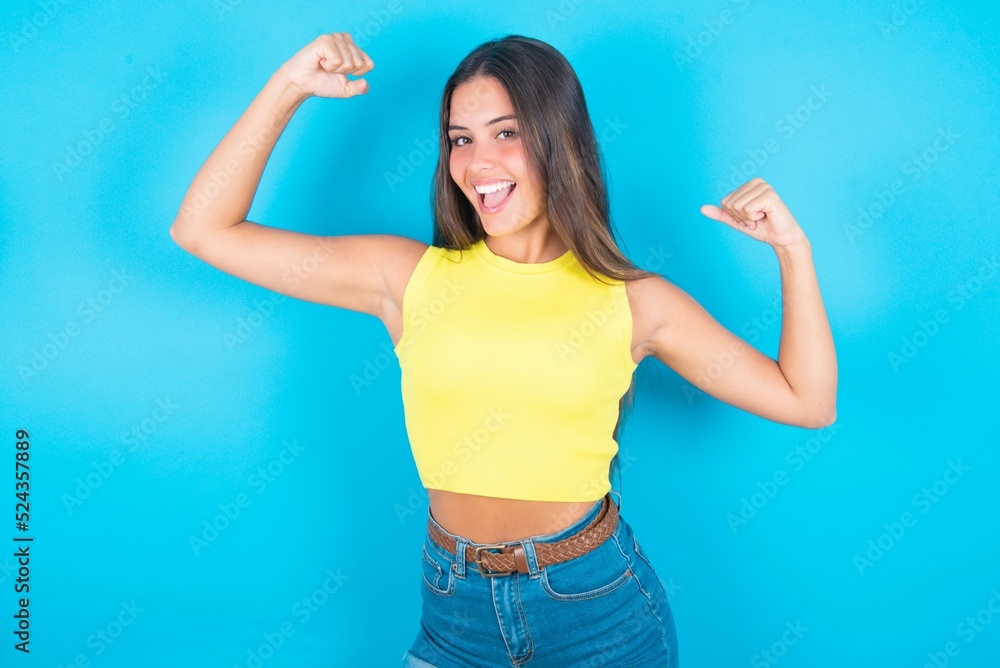 Yes I am winner. Portrait of charming delighted and excited brunette woman wearing yellow tank top over blue raising up fist in triumph and victory smiling achieving success grinning from delight. Yes I am winner. Portrait of charming delighted and excited brunette woman wearing yellow tank top over blue raising up fist in triumph and victory smiling achieving success grinning from delight.