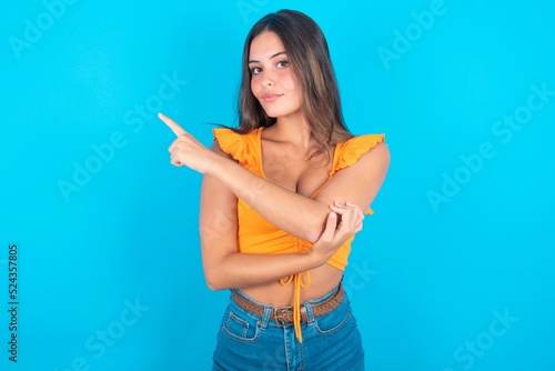 Portrait of brunette woman wearing orange tank top over blue background posing on camera with tricky look  presenting product with index finger. Advertisement concept.