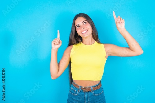 Successful friendly looking brunette woman wearing yellow tank top over blue background exclaiming excitedly, pointing both index fingers up, indicating something.