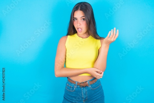 Obraz na plátně Studio shot of frustrated brunette woman wearing yellow tank top over blue background gesturing with raised palm, frowning, being displeased and confused with dumb question