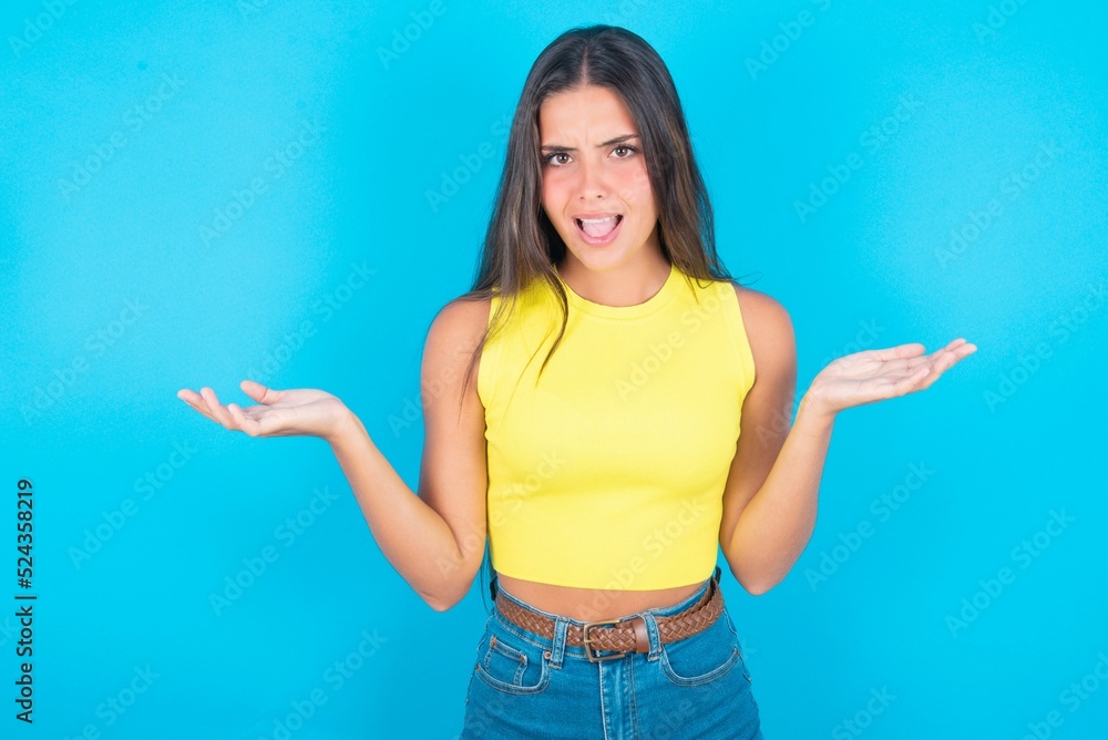 So what? Portrait of arrogant brunette woman wearing yellow tank top over blue background shrugging hands sideways smiling gasping indifferent, telling something obvious. So what? Portrait of arrogant brunette woman wearing yellow tank top over blue background shrugging hands sideways smiling gasping indifferent, telling something obvious.