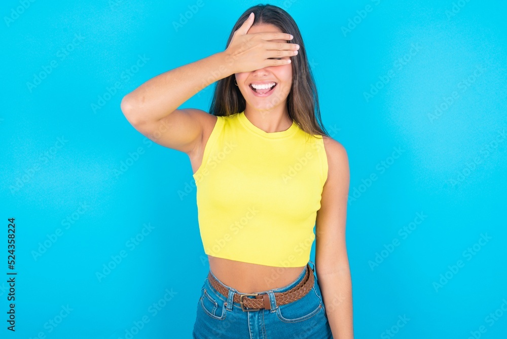 Happy brunette woman wearing yellow tank top over blue background closing eyes with hand going to see surprise prepared by friend standing and smiling in anticipation for something wonderful. Happy brunette woman wearing yellow tank top over blue background closing eyes with hand going to see surprise prepared by friend standing and smiling in anticipation for something wonderful.