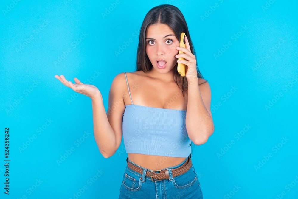 Fototapeta premium beautiful brunette woman wearing blue tank top over blue backgr talking on the phone stressed with hand on face, shocked with shame and surprise face, angry and frustrated. Fear and upset for mistake.