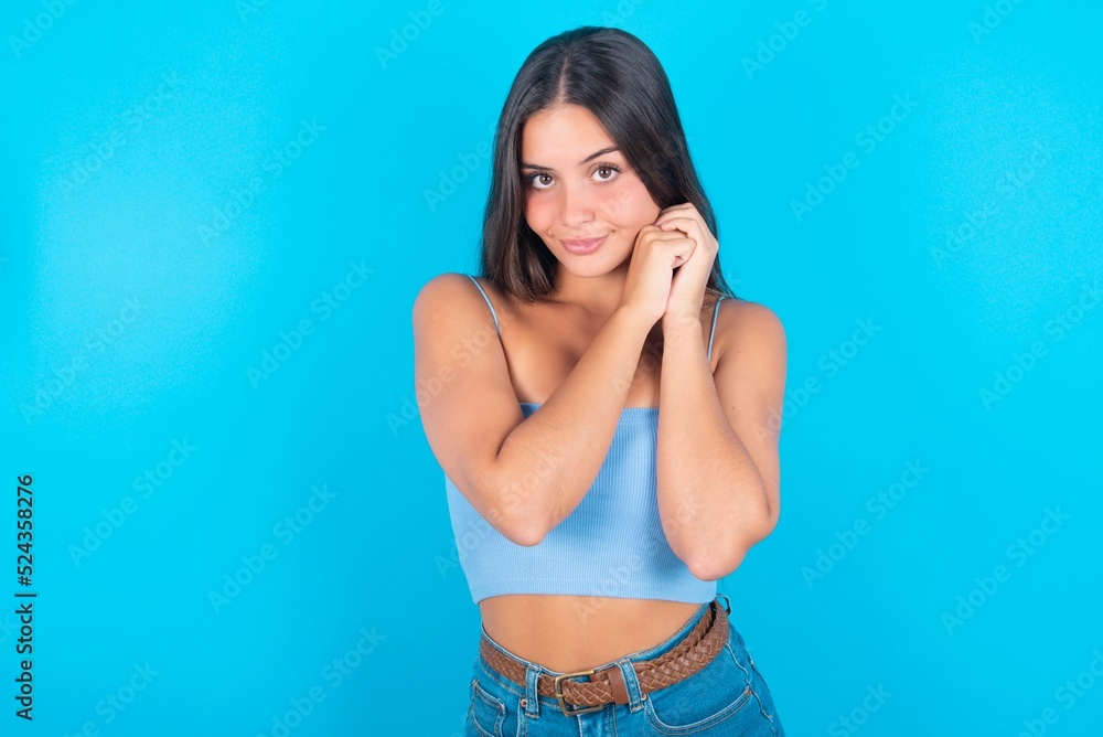 Charming serious beautiful brunette woman wearing blue tank top over blue background keeps hands near face smiles tenderly at camera Charming serious beautiful brunette woman wearing blue tank top over blue background keeps hands near face smiles tenderly at camera
