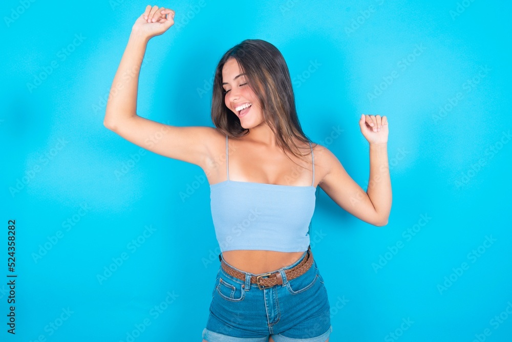 Photo of upbeat beautiful brunette woman wearing blue tank top over blue background has fun and dances carefree wear being in perfect mood makes movements. Spends free time on disco party Photo of upbeat beautiful brunette woman wearing blue tank top over blue background has fun and dances carefree wear being in perfect mood makes movements. Spends free time on disco party