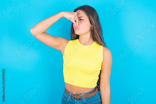 Displeased brunette woman wearing yellow tank top over blue background plugs nose as smells something stink and unpleasant, feels aversion, hates disgusting scent.