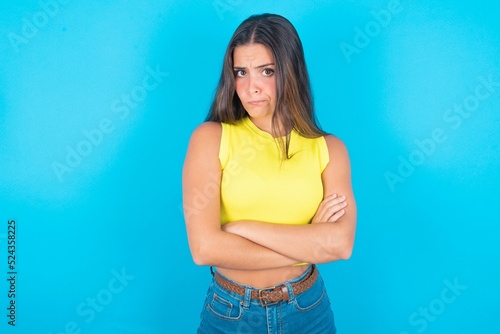Picture of angry brunette woman wearing yellow tank top over blue background crossing arms. Looking at camera with disappointed expression. © Jihan