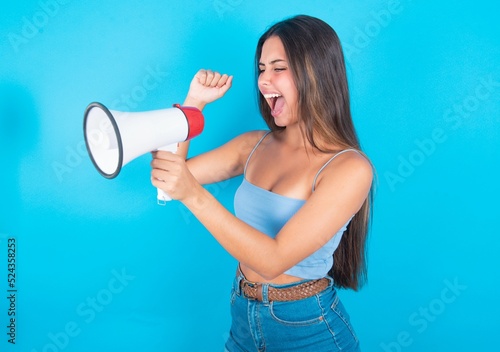 beautiful brunette woman wearing blue tank top over blue background communicates shouting loud holding a megaphone, expressing success and positive concept, idea for marketing or sales.
