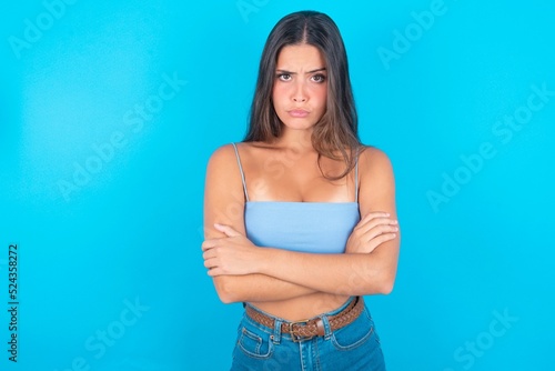 Gloomy dissatisfied beautiful brunette woman wearing blue tank top over blue background looks with miserable expression at camera from under forehead, makes unhappy grimace