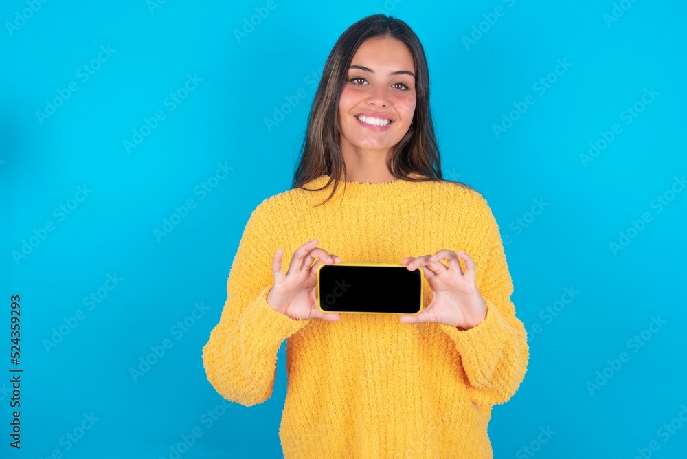 Cheerful cheery content beautiful brunette woman wearing yellow sweater over blue background holding in hands device hobby smm post blog