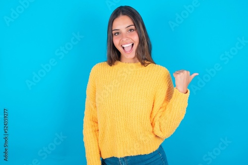 Impressed beautiful brunette woman wearing yellow sweater over blue background point back empty space