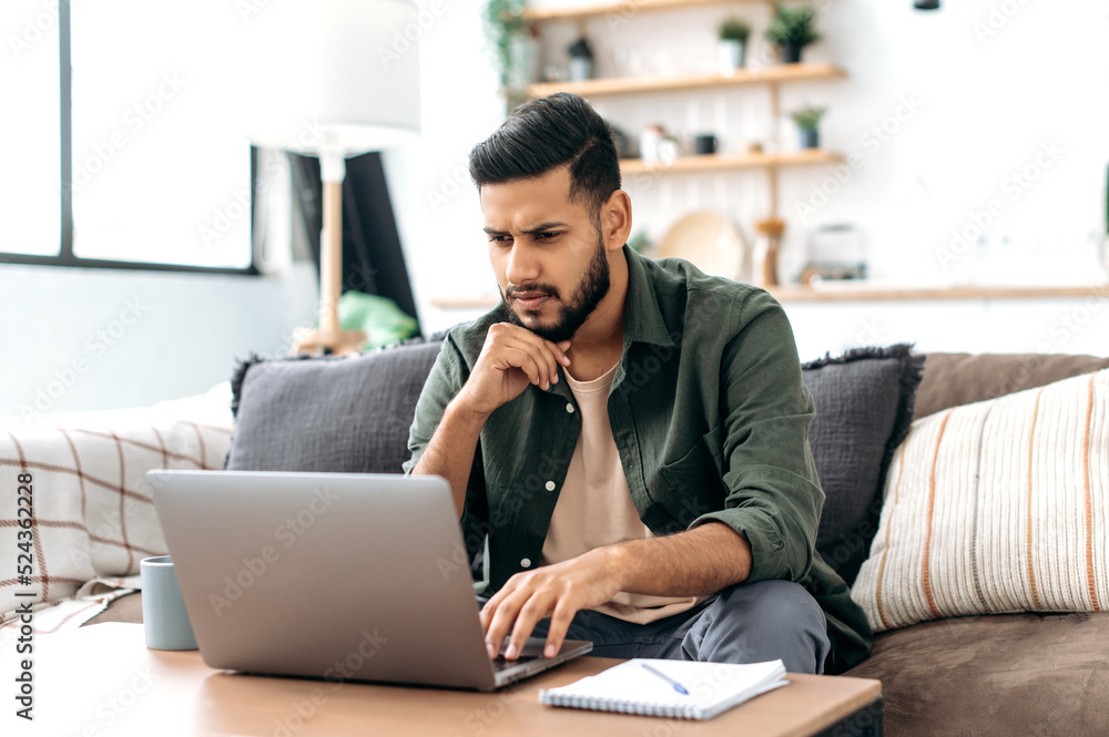 Focused smart arabian or indian young man in stylish casual clothes, freelancer, web designer, sitting in the living room on the sofa, working on a laptop on a project, looking at the screen intently Focused smart arabian or indian young man in stylish casual clothes, freelancer, web designer, sitting in the living room on the sofa, working on a laptop on a project, looking at the screen intently