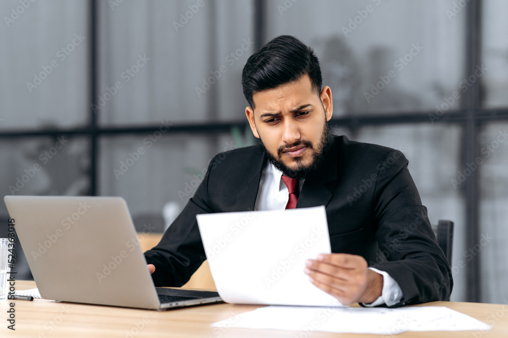 Frustrated tired indian or arabian business man, sitting upset at work desk with laptop in modern office, works with documents, makes a financial report, analyzes the result of work, feels overworked Frustrated tired indian or arabian business man, sitting upset at work desk with laptop in modern office, works with documents, makes a financial report, analyzes the result of work, feels overworked