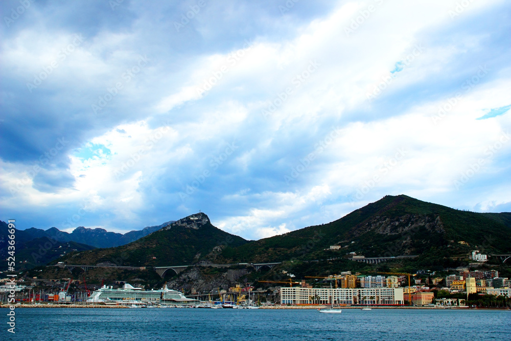Interesting view of the Salerno coast with crests of impressive mountains standing out from the densely populated area crossed by a tall bridge and skimming the blue sea under a cloudy summer day