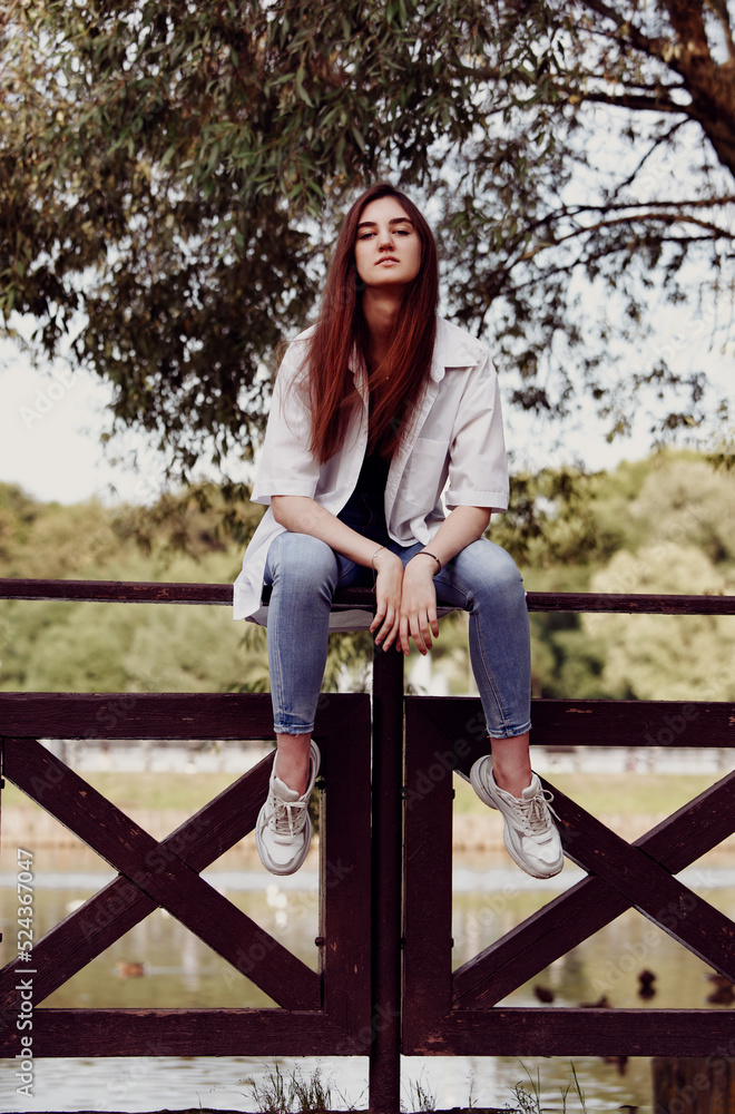 Tthinking teen casual woman in blue jeans sitting on the fence outdoors on summer green trees background. Closeup portrait in sunny day in the park Tthinking teen casual woman in blue jeans sitting on the fence outdoors on summer green trees background. Closeup portrait in sunny day in the park