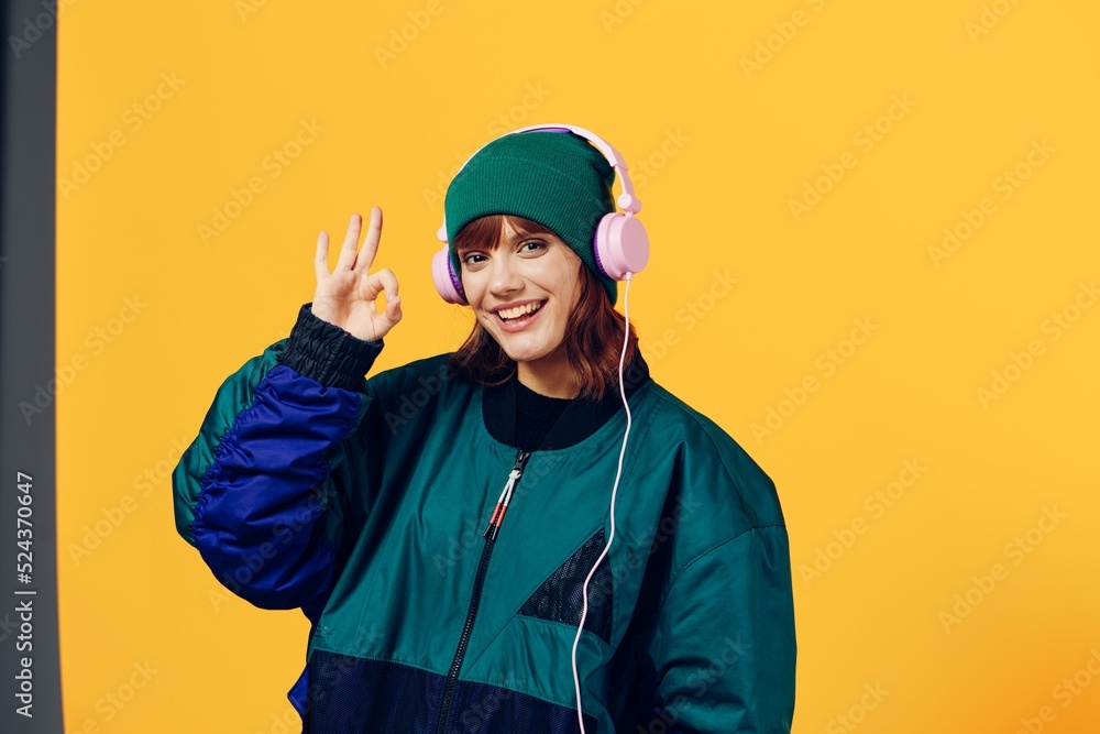 a joyful woman in a green jacket and a hat in the style of the 90s enjoys music standing in pink headphones on a yellow background showing the ok sign while looking at the camera