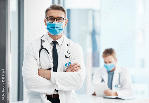 Doctor  physician or healthcare professional with covid face mask in a hospital for medical health insurance background. Innovation  leadership and excellence male gp portrait with his arms crossed