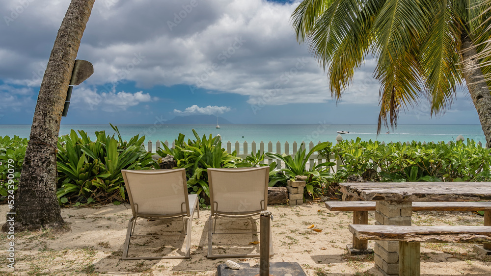 Naklejka premium A relaxation area on a tropical beach. On the sand under the palm trees there are sun loungers, wooden benches, a table. Ahead, behind the fence - turquoise ocean, blue sky with clouds. Seychelles. 