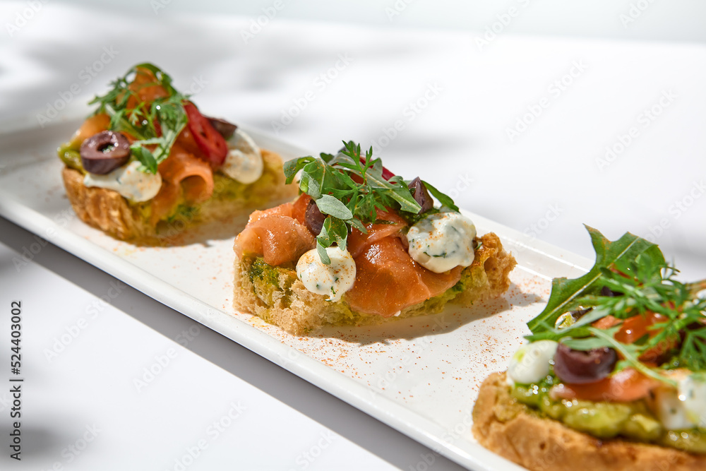 Aesthetic composition with salmon bruschetta on white background with shadows from flowers. Italian bruschetta with salmon, avocado, cheese and olives on fine dining in summer. Elegant menu concept.