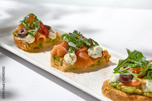 Aesthetic composition with salmon bruschetta on white background with shadows from flowers. Italian bruschetta with salmon, avocado, cheese and olives on fine dining in summer. Elegant menu concept.