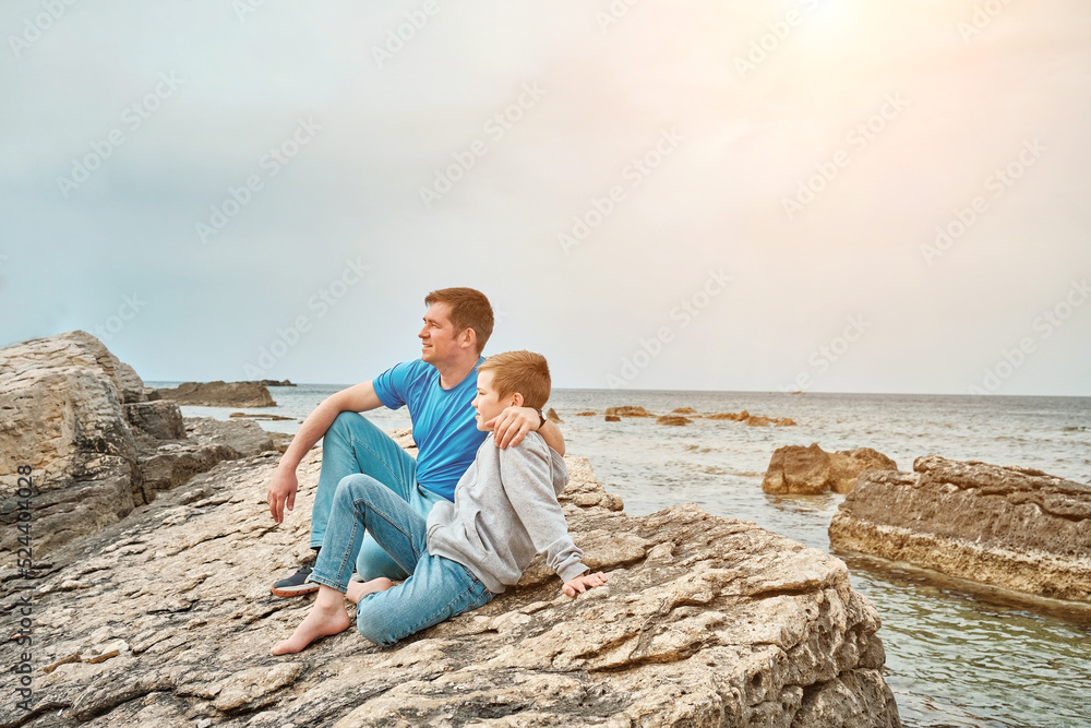 Happy family, father and son bonding, sitting on stone by the sea looking at view enjoying summer vacation. Togetherness Friendly concept	
