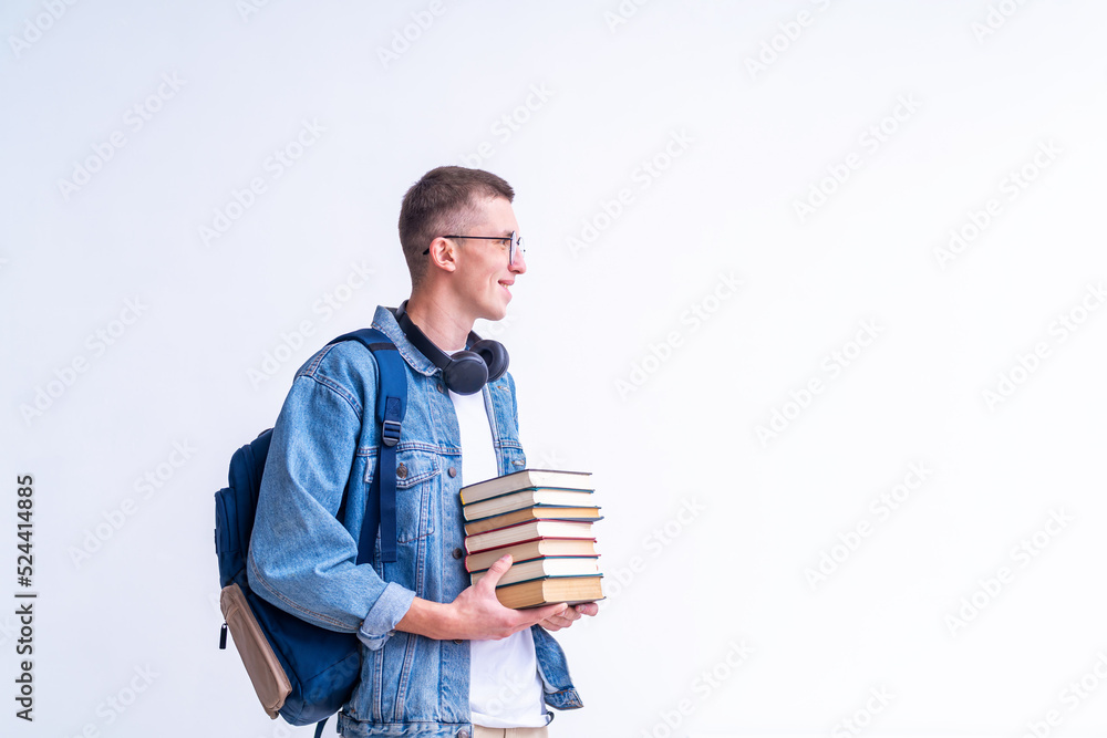 cheerful teenage with glasses, headphones, backpack and stack books in his hands stands against white wall. the smiling student looks away. emotional portrait beautiful teenage boy returning to school cheerful teenage with glasses, headphones, backpack and stack books in his hands stands against white wall. the smiling student looks away. emotional portrait beautiful teenage boy returning to school