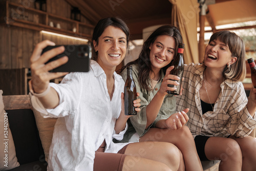 Three cheerful young caucasian ladies with beer take photo on smartphone spending leisure time at party. Brunettes smile, wear casual clothes. Happy weekend concept.