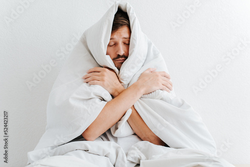 Adult frozen man wrapped in a blanket and hugging himself while sitting on a bed indoors. Lonely guy with closed eyes warming in a cold house photo