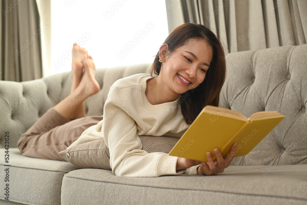 Smiling young woman lying on couch and reading book, enjoy stress free peaceful mood in cozy winter or autumn weekend Smiling young woman lying on couch and reading book, enjoy stress free peaceful mood in cozy winter or autumn weekend