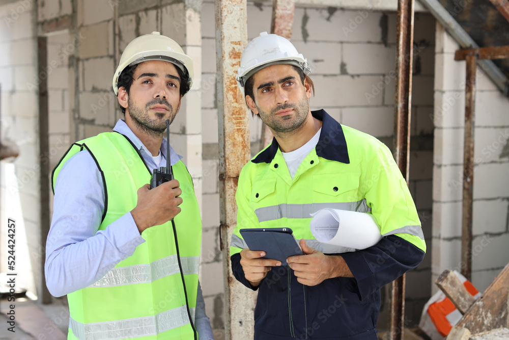Two technician civil engineer or specialist inspector discussing, brainstorm and planing work with tablet, blueprint and walkie talkie radio together at Industrial building site. Construction concept Two technician civil engineer or specialist inspector discussing, brainstorm and planing work with tablet, blueprint and walkie talkie radio together at Industrial building site. Construction concept