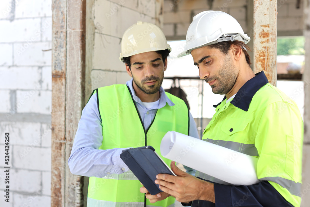 Two technician civil engineer or specialist inspector discussing, brainstorm and planing work with tablet, blueprint and walkie talkie radio together at Industrial building site. Construction concept Two technician civil engineer or specialist inspector discussing, brainstorm and planing work with tablet, blueprint and walkie talkie radio together at Industrial building site. Construction concept