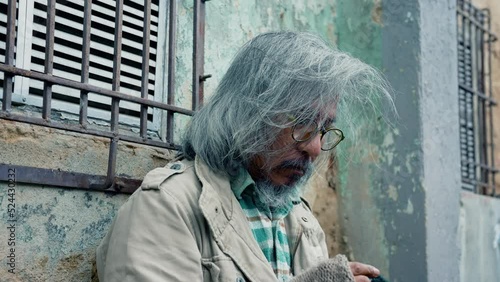 4K, Close Up A homeless man in a dirty shirt with long hair and glasses sits hopelessly against an old wall, waiting for help from passersby. A man without a home lives in poverty on the streets. photo