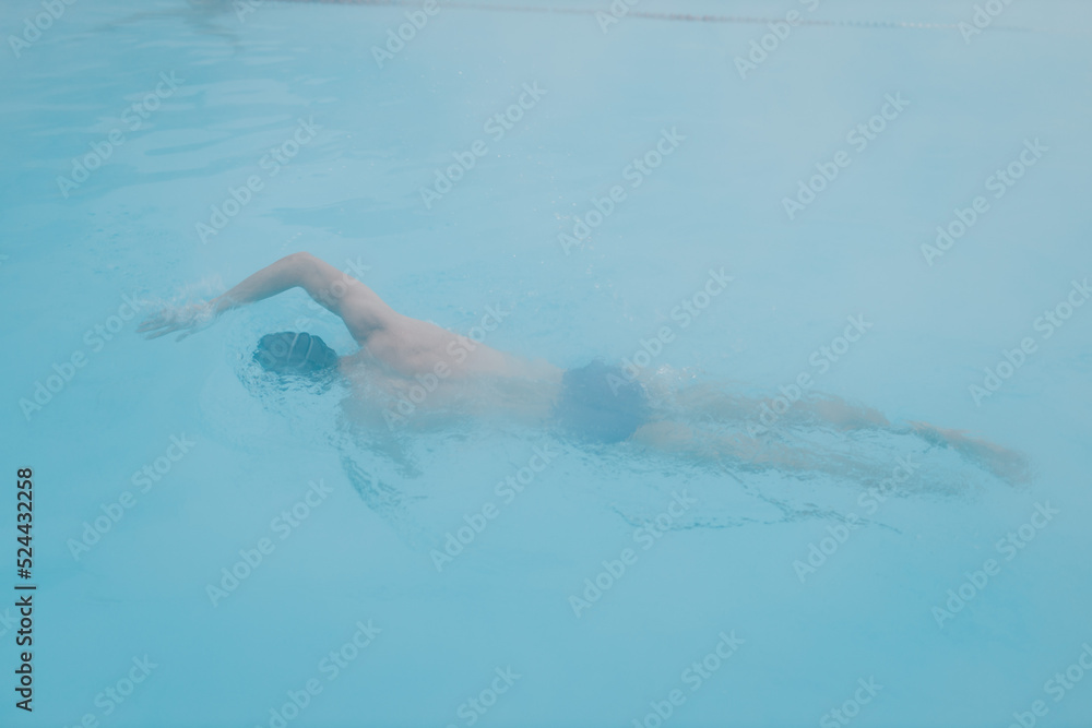 Fit swimmer male training swim crawl in open winter swimming pool with fog. Geothermal outdoor spa health concept.