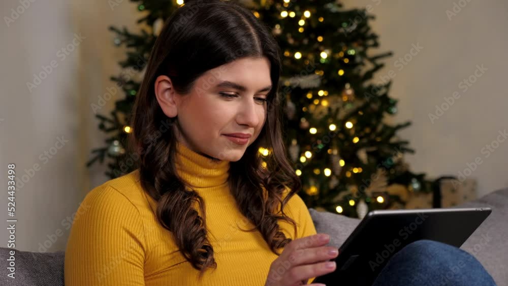 Close up smiling woman buys New Year gifts on internet using tablet on background Christmas tree. Positive female uses portable device online chatting sitting on couch at cozy home. Winter holidays Close up smiling woman buys New Year gifts on internet using tablet on background Christmas tree. Positive female uses portable device online chatting sitting on couch at cozy home. Winter holidays