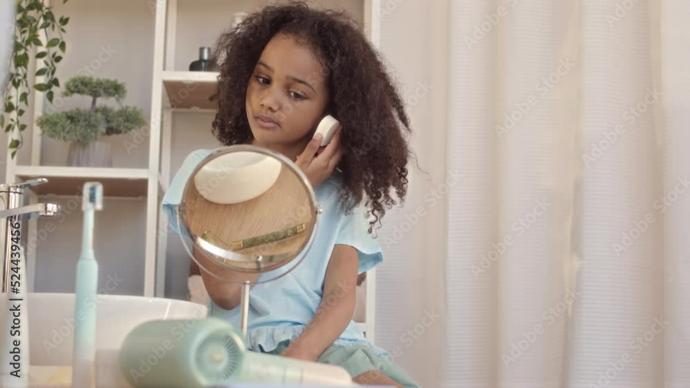 Medium slowmo of pretty Biracial 6 year old girl using sponge while doing her makeup, looking at herself in mirror at bathroom Medium slowmo of pretty Biracial 6 year old girl using sponge while doing her makeup, looking at herself in mirror at bathroom