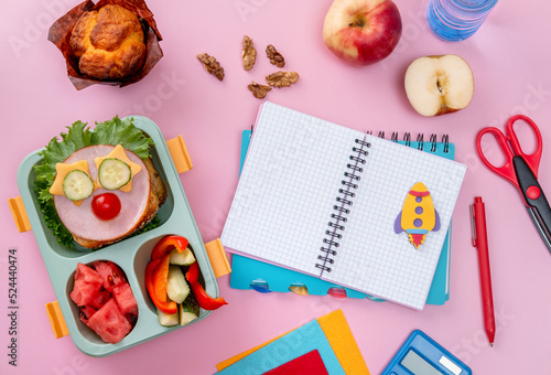 School lunch box for kids with food in the form of funny faces. School lunch box with sandwich, vegetables, water and stationery on table. Healthy eating habits concept. Back to school concept photo