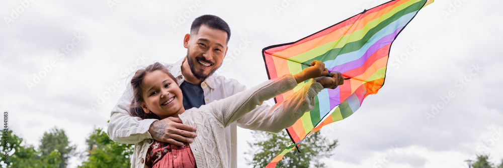 Smiling asian man hugging preteen daughter with flying kite in park, banner. Smiling asian man hugging preteen daughter with flying kite in park, banner.